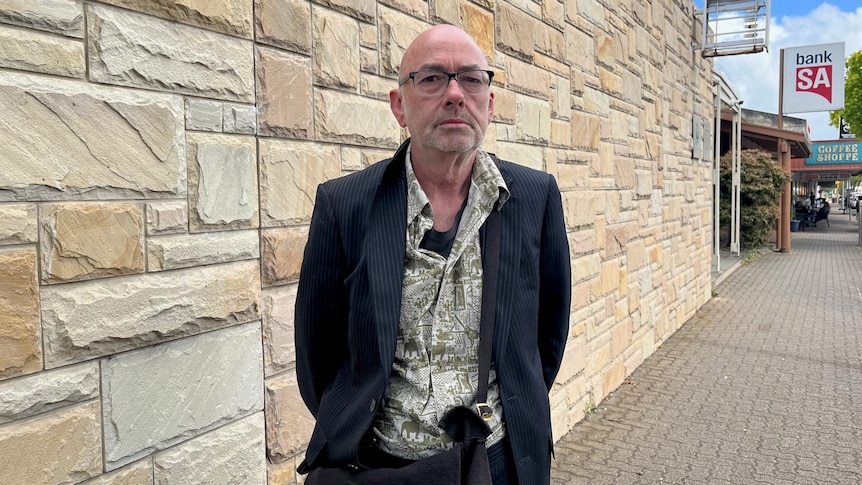 A bald man wearing a suit and floral shirt standing outside a sandstone wall