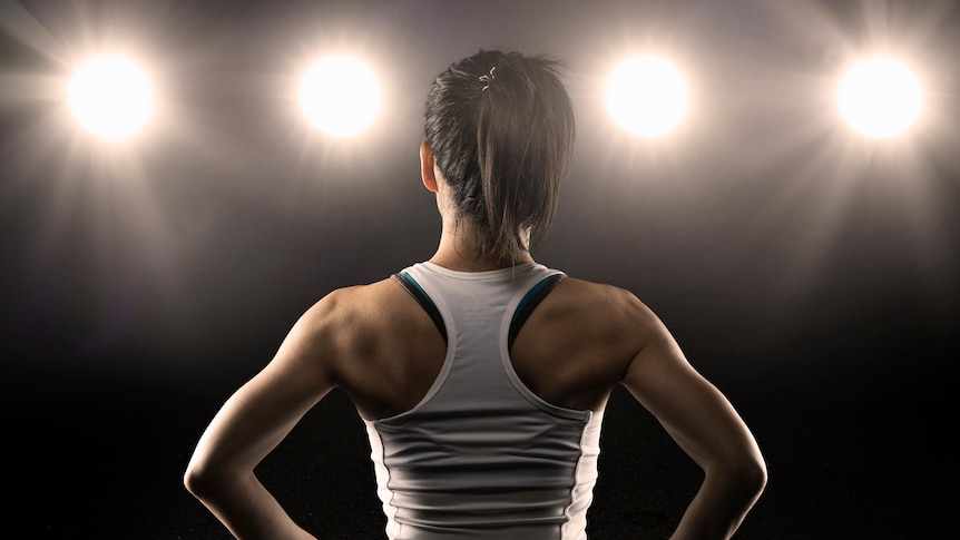 A woman in activewear with her hands on her hips, seen from behind with spotlights shining from in front of her.