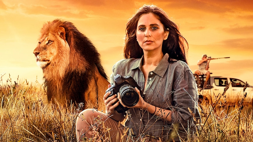 Woman holding camera with a lion and game hunter in the background.