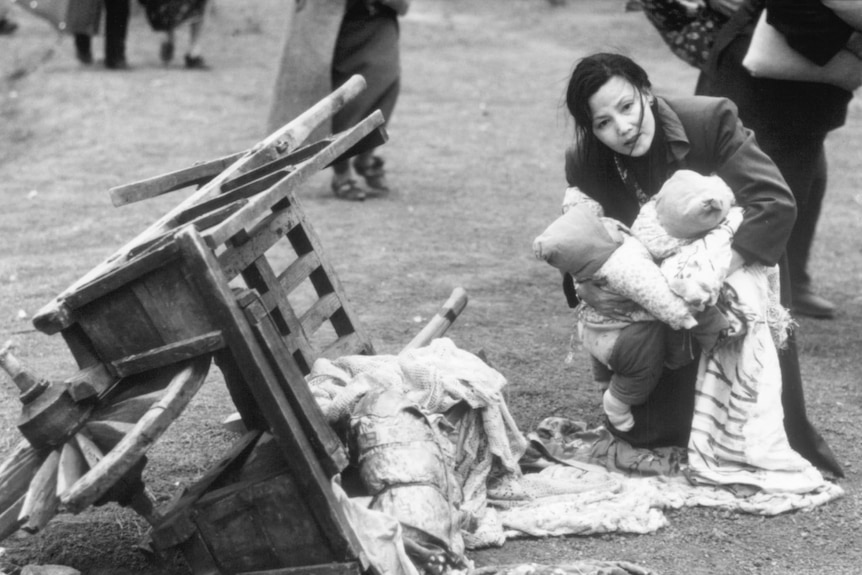 Black and white film still of Kieu Chinh kneeling in the street, holding two babies 