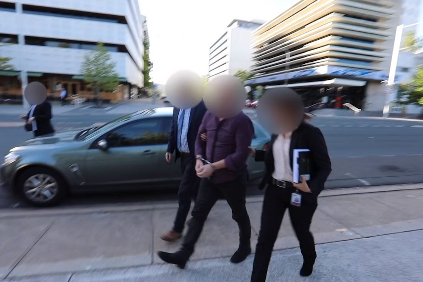 A man, whose face is blurred is walked down the street in handcuffs with two officers.