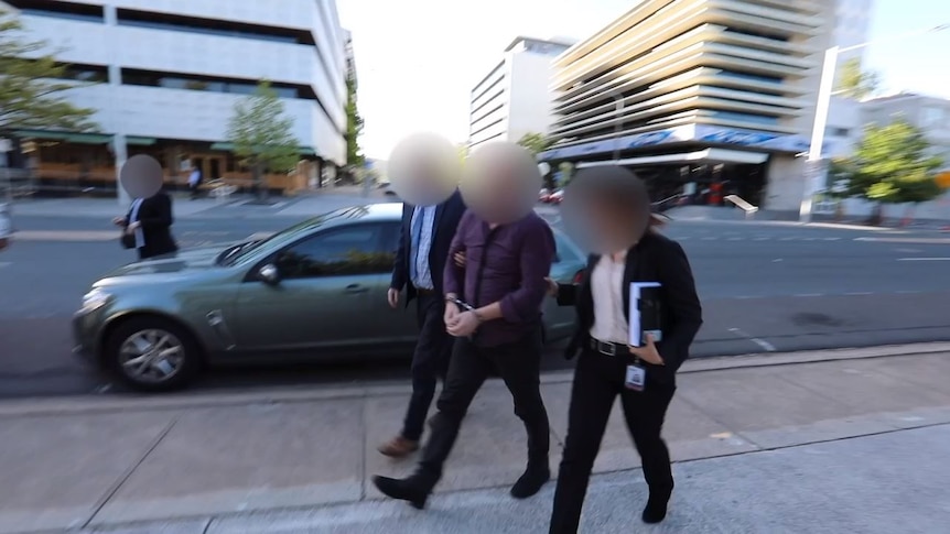 A man, whose face is blurred is walked down the street in handcuffs with two officers.