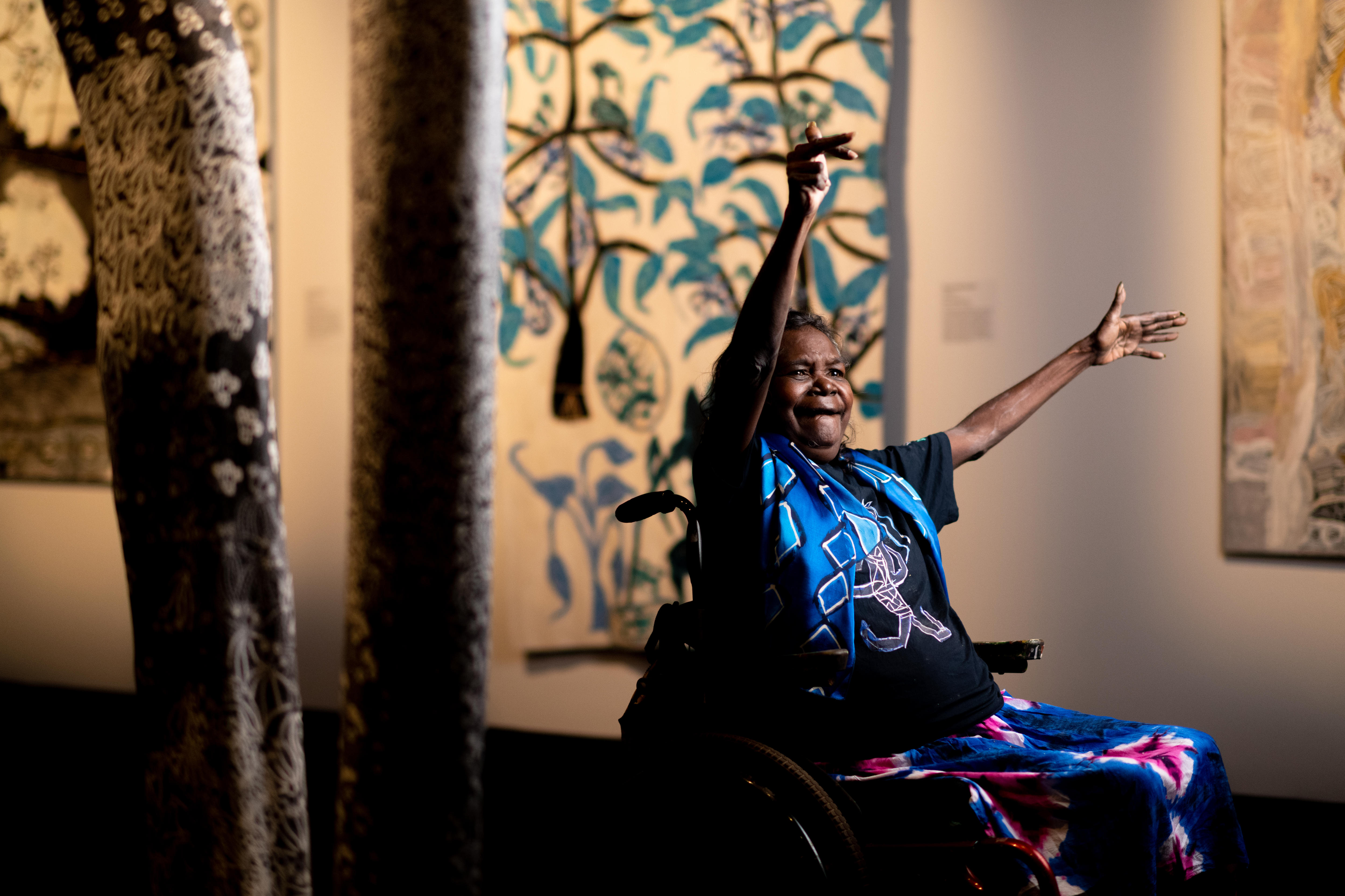 Artist Dhambit Munungurr sits in her wheelchair, smiling, with her hands raised in celebration.