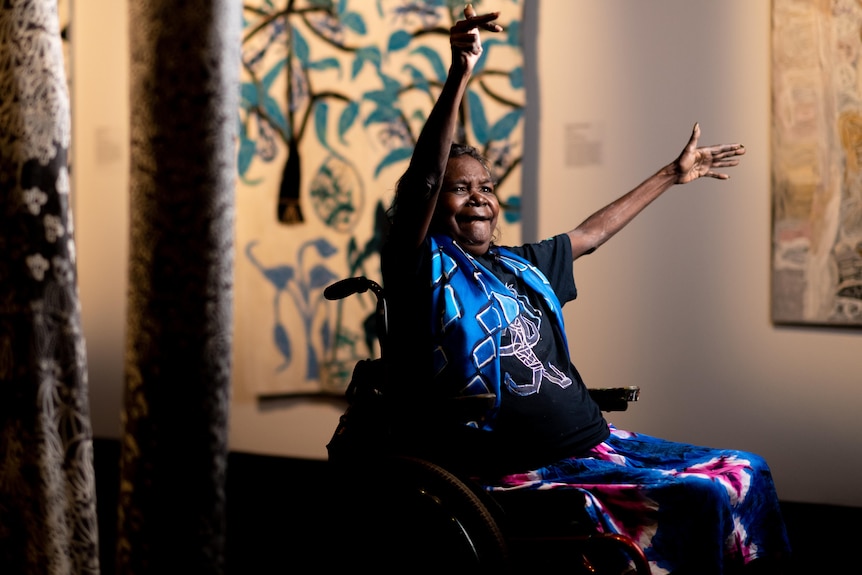 Die Künstlerin Dhambit Munungurr sitzt lächelnd in ihrem Rollstuhl, die Hände zur Feier erhoben.
