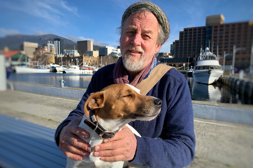 A man holds a dog in front of a city and port landscape.
