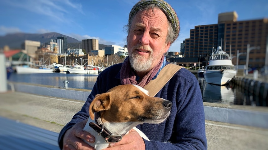 A man holds a dog in front of a city and port landscape.