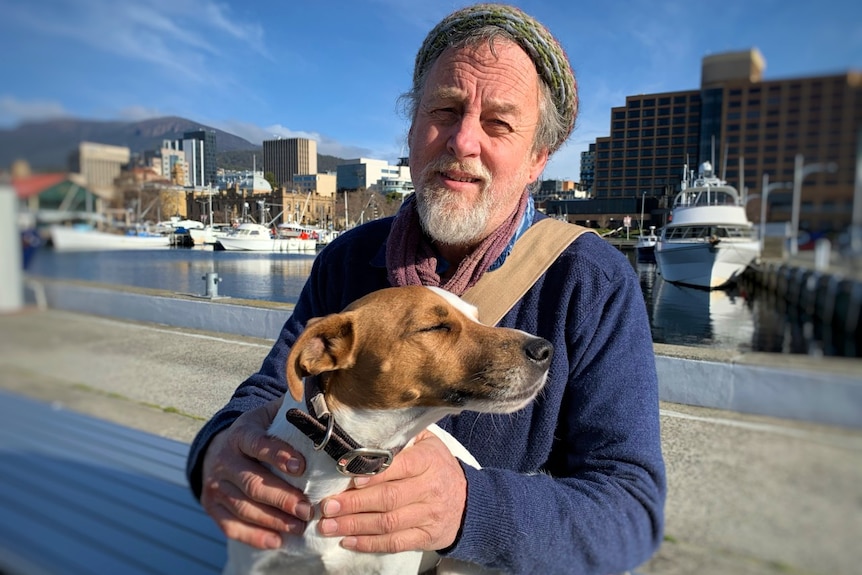 A man holds a dog in front of a city and port landscape.