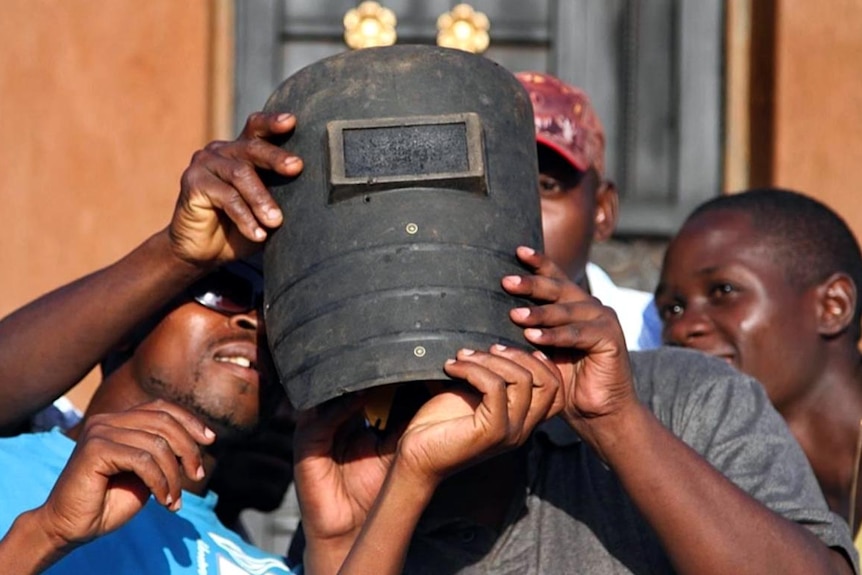 A group of men in Uganda watch a hybrid solar eclipse through a welder's mask.