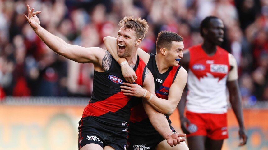 A male AFL player smiles and raises his right hand as he is hugged around his neck by a teammate in celebration.