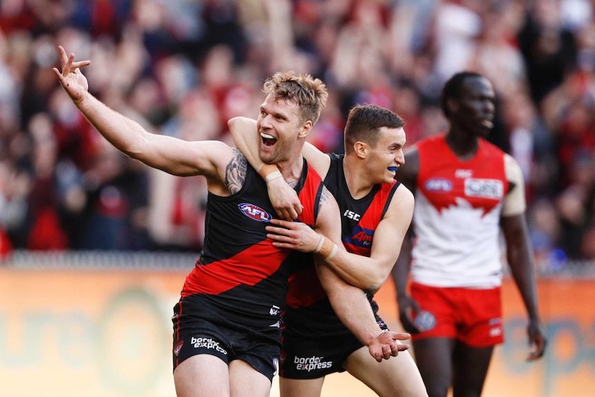 A male AFL player smiles and raises his right hand as he is hugged around his neck by a teammate in celebration.