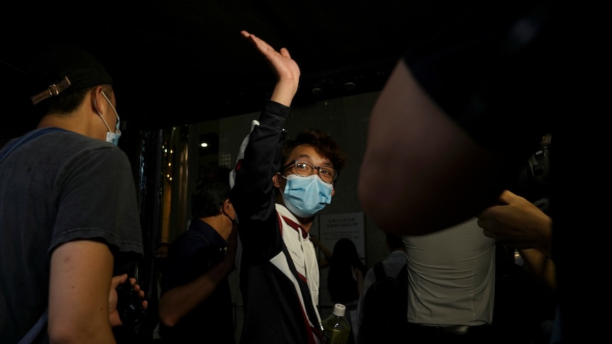 A young Chinese man in white shirt and blue face mask waves to crowd as he walks in dark corridor surrounded by two men.