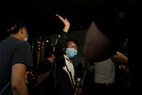 A young Chinese man in white shirt and blue face mask waves to crowd as he walks in dark corridor surrounded by two men.