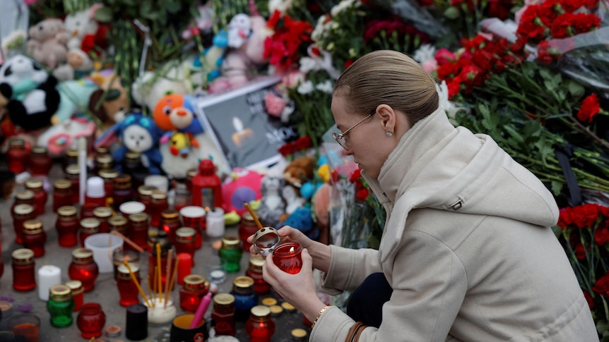 A woman crouches down to the ground to light a candle surrounded by other candles, flowers and plush toys.
