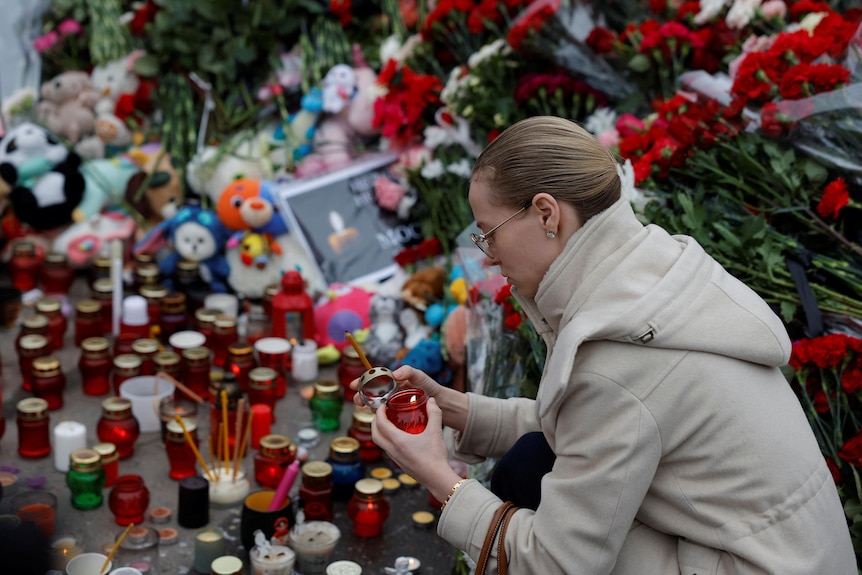 A woman crouches down to the ground to light a candle surrounded by other candles, flowers and plush toys.