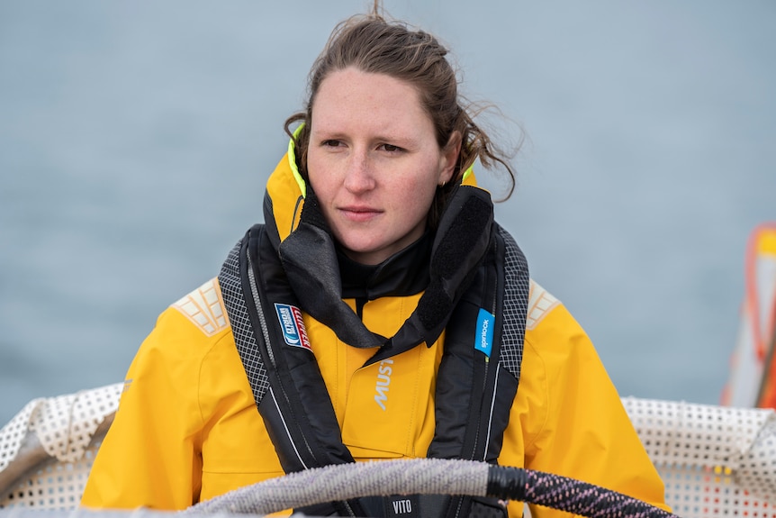 Hannah Brewis stares out in front of her wearing a yellow jacket and with the ocean behind her. 