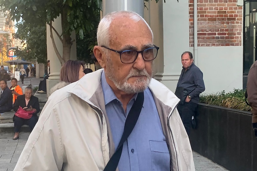 A head shot of an older man with grey balding hair and a beard, in spectacles, a beige jacket and a blue shirt, outside court.