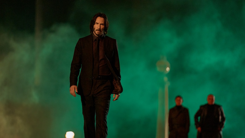 Tall white man with black shoulder-length hair wears a black suit and walks on a street with green smoke rising behind him.