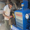Mintu, an Indian Sikh, wears a beige top and khaki pants while he pours water into a vintage blue Ford tractor engine.