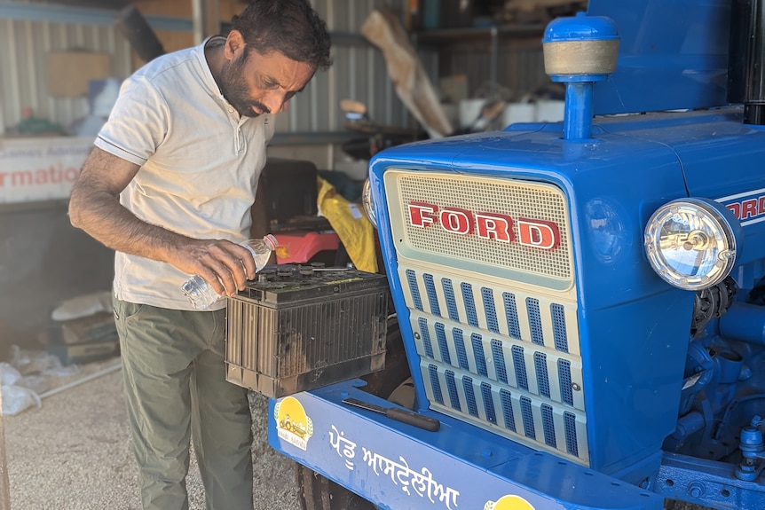 Mintu, a Sikh Indian man, wears a beige top and khaki pants as he pours water into a vintage blue Ford tractor engine.