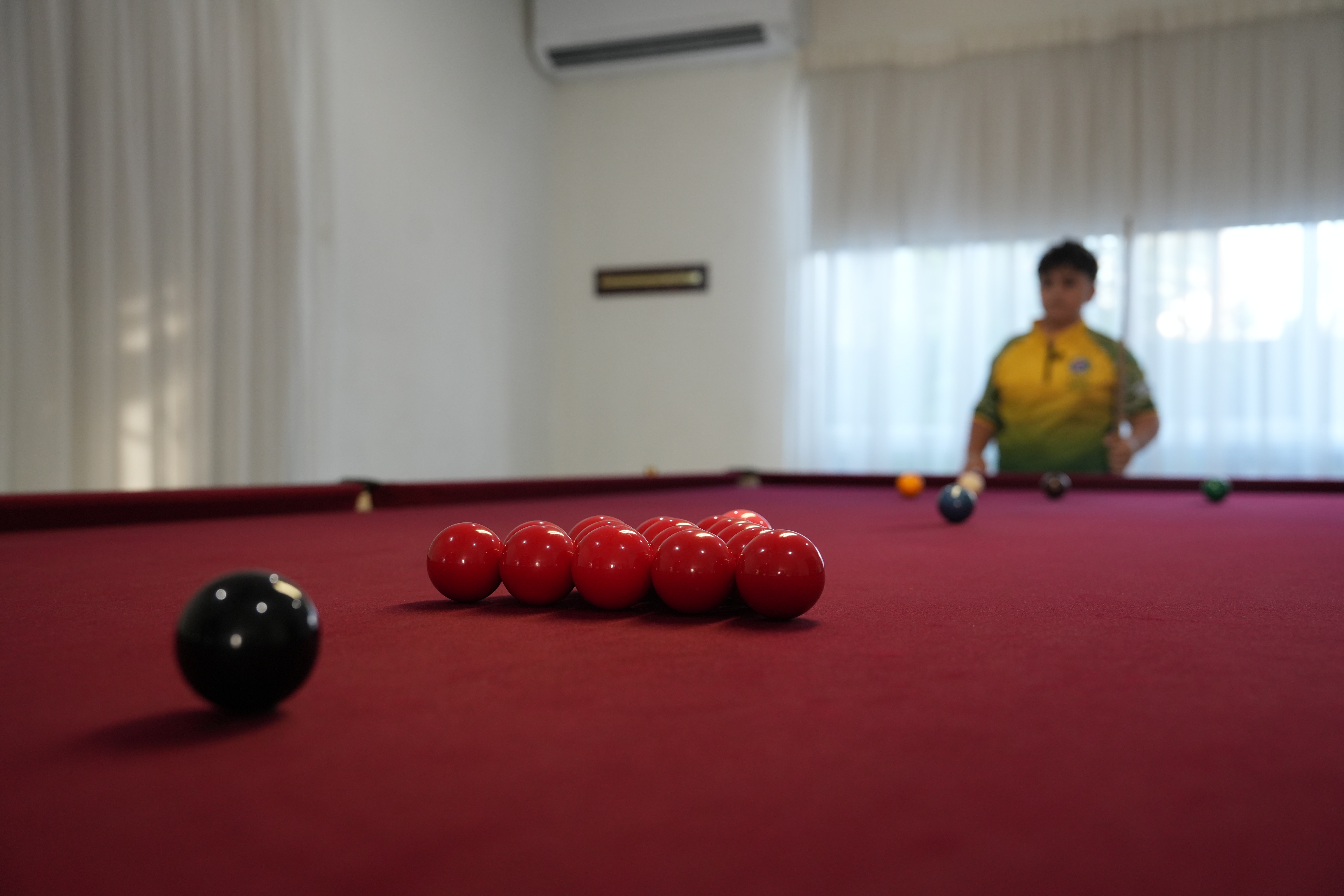 A close-up of red balls lined up on a snooker table with a young boy, Angelo Tsakaris, standing in the background.