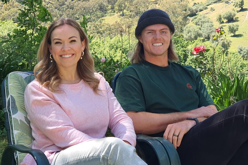 A woman wearing a pink jumper sits next to a man in a green tshirt and blue beanie, both are smiling