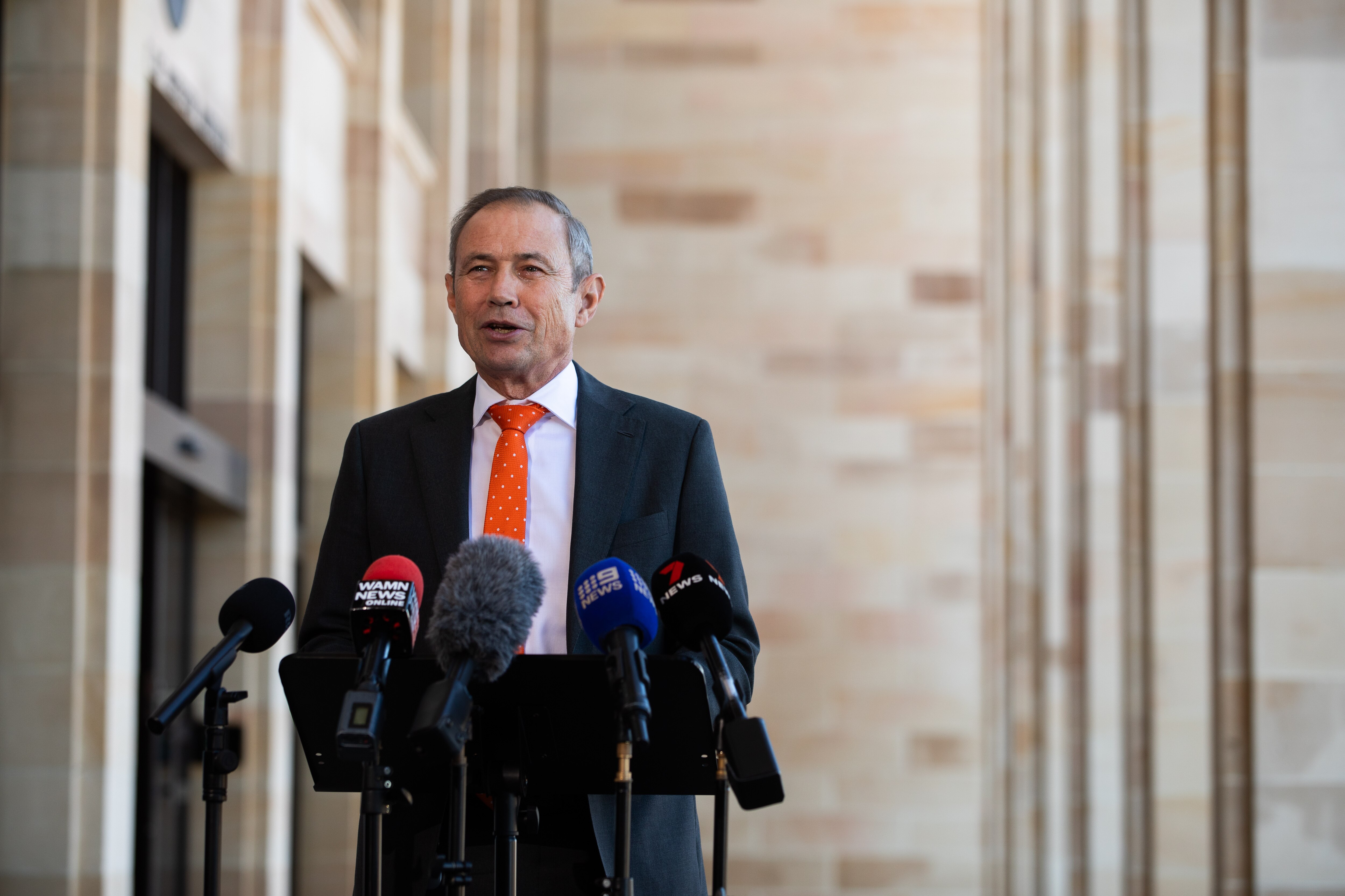 Roger Cook falando do lado de fora de um prédio em frente a microfones.