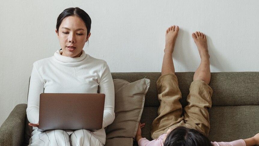 A woman sits on the couch using a laptop as her child lies next to her.