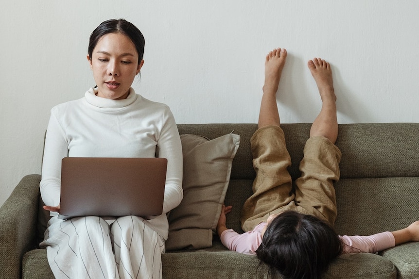 A woman sits on the couch using a laptop as her child lies next to her.