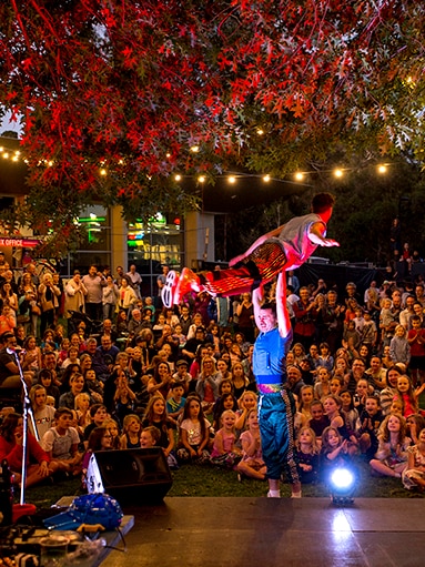 An performer holds another aloft on a stage in front of crowd of children and adults.