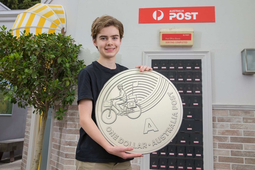 A boy holding a large novelty sized one-dollar coin featuring the design of an Australia Post worker on a bicycle.