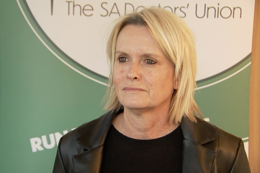 A woman in a dark jacket looking stern in front of a banner that says the SA doctors' union