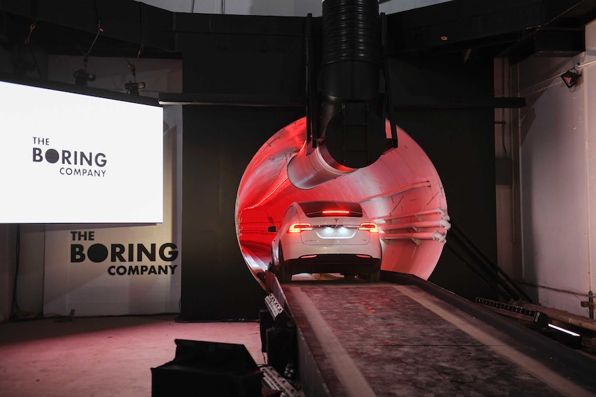 A white Tesla vehicle enters a red-lit tunnel, with white signs reading, "the Boring Company", to the left.