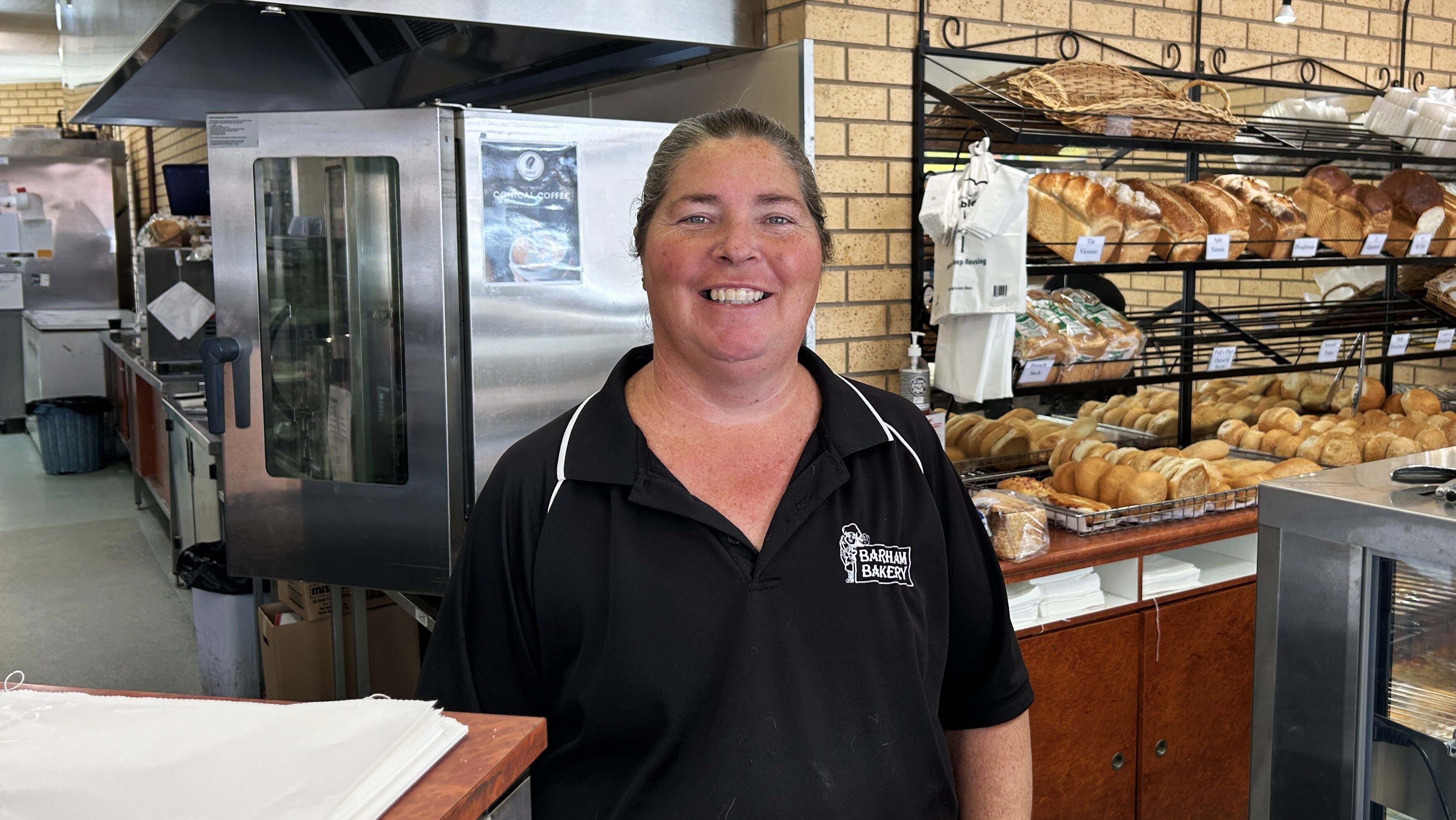 A woman in black polo shirt emblazoned Barham Bakery smiles behind the shop counter.