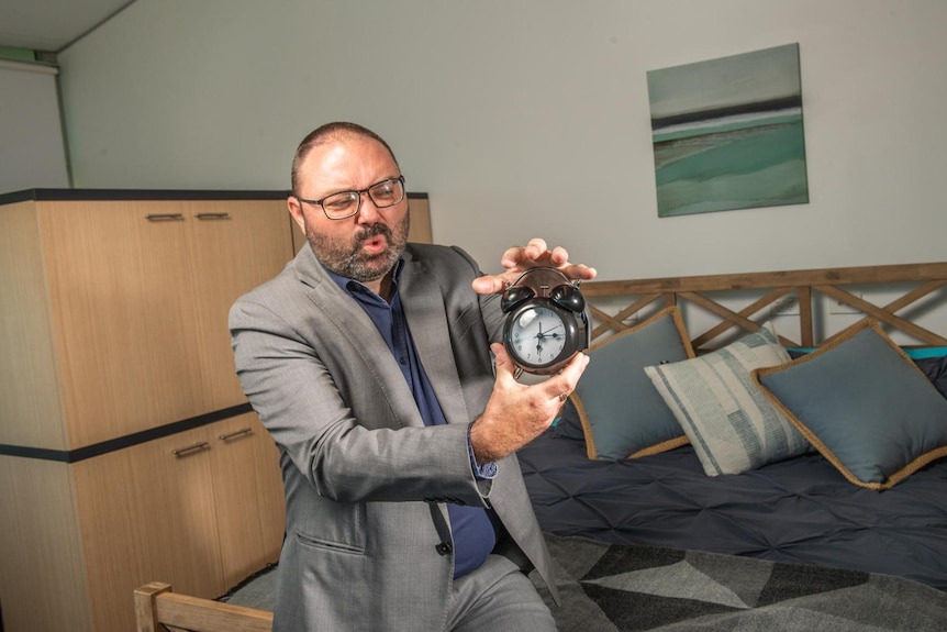 A man holds an alarm clock with a frustrated expression