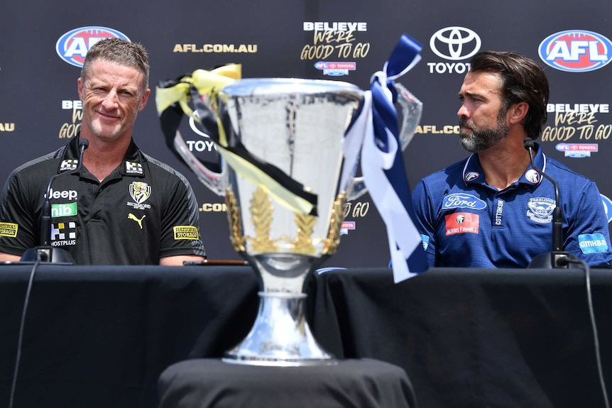 Damien Hardwick smiles while Chris Scott looks at him. Between them sits the AFL premiership cup.