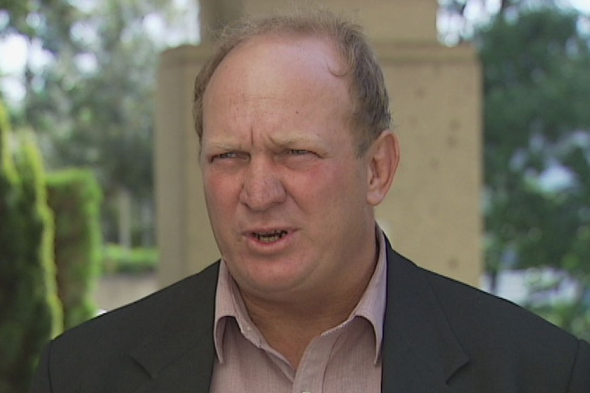 A man with receding hair, wearing a dark blazer, speaking to the media.