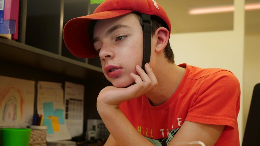 Cooper Sinclair, red t-shirt, red padded cap with strap, brown hair, sitting and concentrating with his hand under his chin.