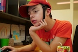Cooper Sinclair, red t-shirt, red padded cap with strap, brown hair, sitting and concentrating with his hand under his chin.