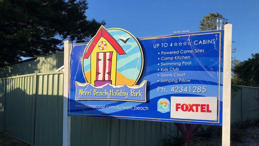 A blue sign to the Werri Beach Holiday Park at Gerringong stands in front of a green fence