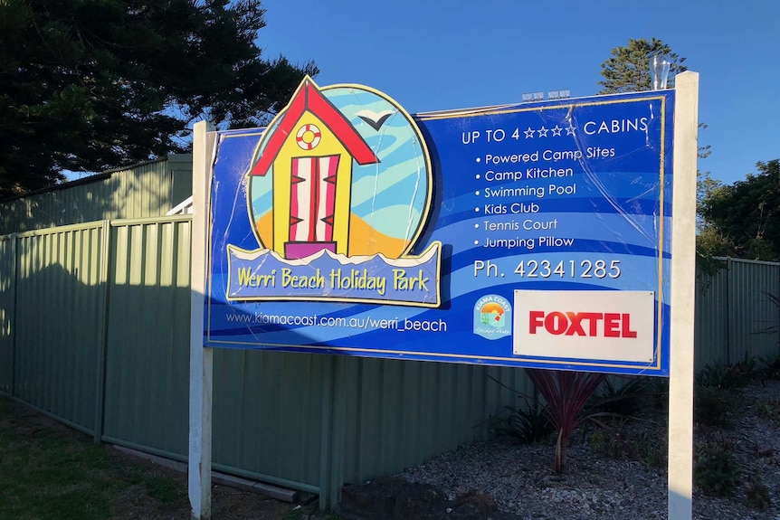 A blue sign to the Werri Beach Holiday Park at Gerringong stands in front of a green fence