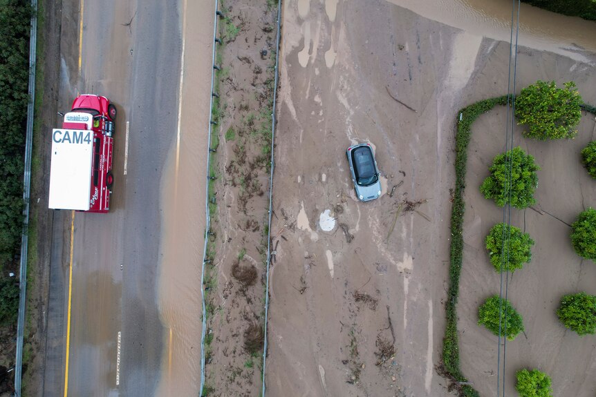 Birds eye view of a car stuck in mud and flood water on a road with a truck driving past