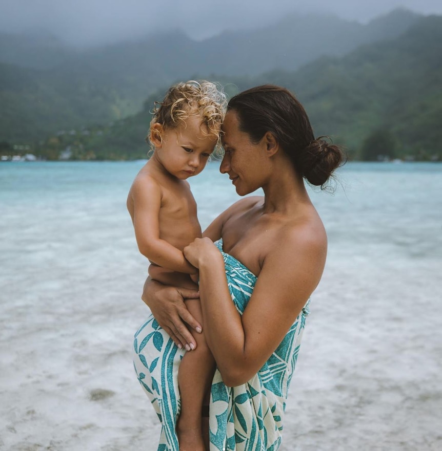 Woman wrapped in lavalava stands on clear water beach and mountains in background, holding toddler on pregnant belly. 