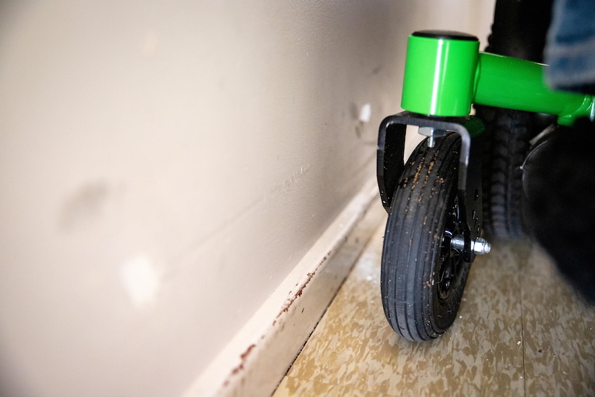 Close-up of the wheel of a wheelchair next to a dented interior wall with a scuffed skirting board.