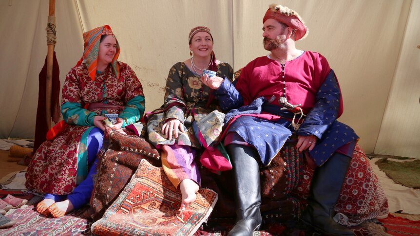 Two women and a man in colourful costumes sit on a carpet-covered ottoman inside a canvas tent.