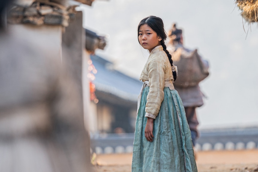 A young Korean girl in traditional dress  stands on a dirt road that leads to the ocean. 