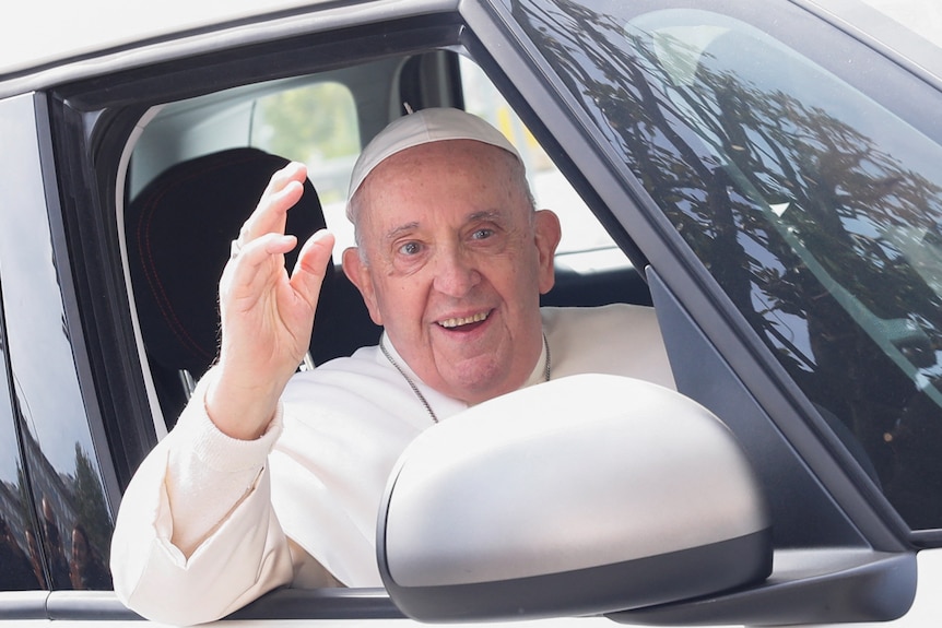 An elderly man in a white robe with a white skull cap smiles as he waves from a car window.