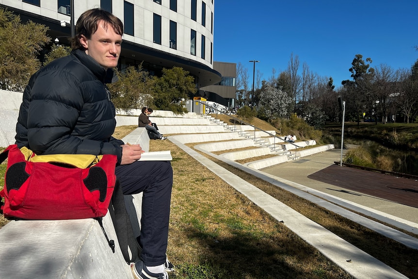 A young person wearing a dark blue puffer jacket with a red bag sits on steps at university with a notebook in their hands.