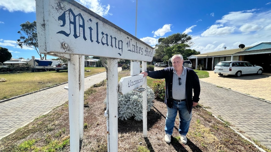A man with grey hair, a navy cardigan and jeans leans against a motel sign reading 'Milang Lakes Motel'