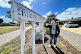 A man with grey hair, a navy cardigan and jeans leans against a motel sign reading 'Milang Lakes Motel'