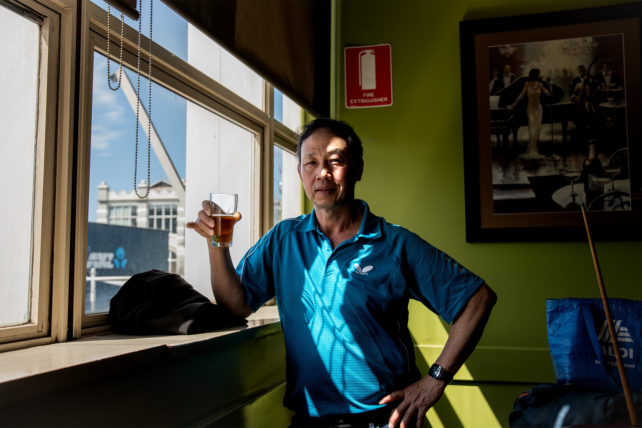 A man wearing a blue polo shirt stands leaning on a window sill, holding a glass of beer in his hand.
